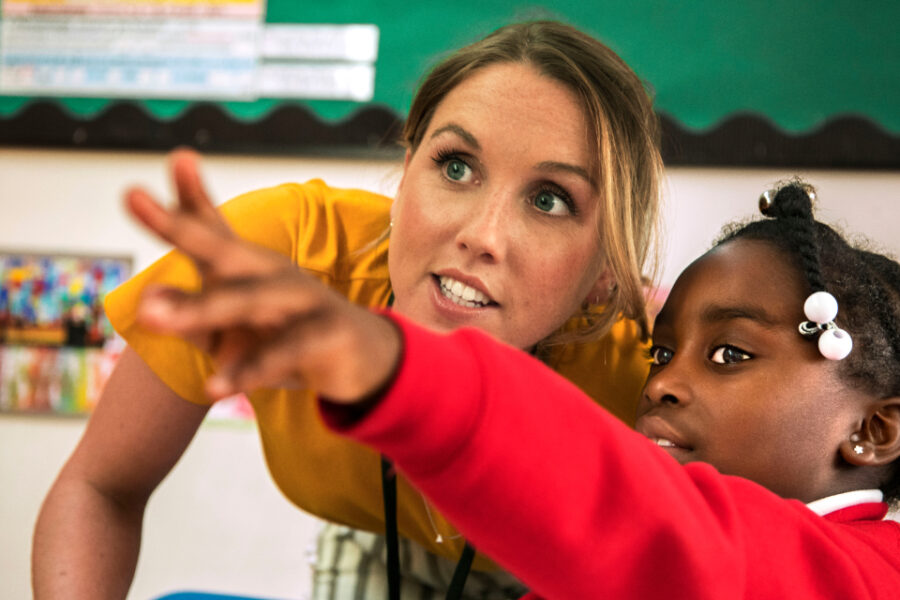 A close up photo of a teacher working together with a student who is pointing the the whiteboard at the front of the classroom.